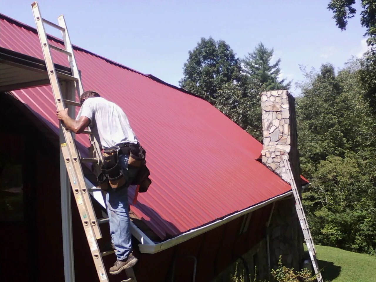 Roofer on a ladder beside a red metal roof and stone chimney — Soffit & Fascia Repair by Roofing Brothers in Wayland